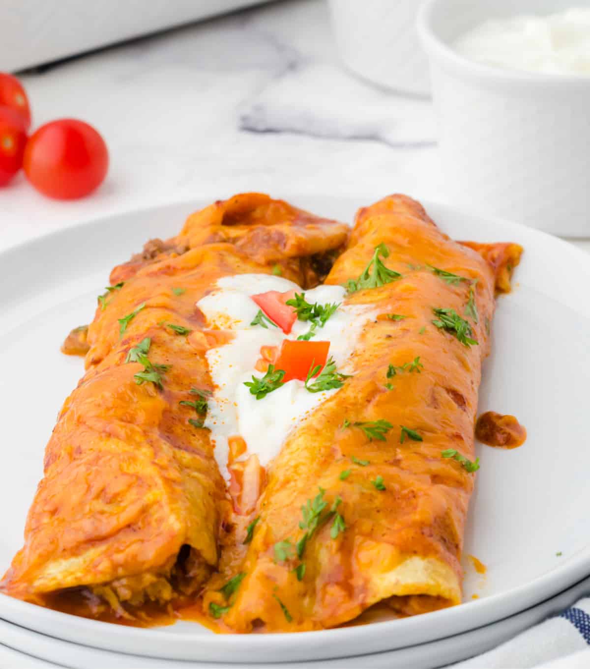 Two beef enchiladas on a white plate with sour cream in the middle and a few diced tomatoes on top.