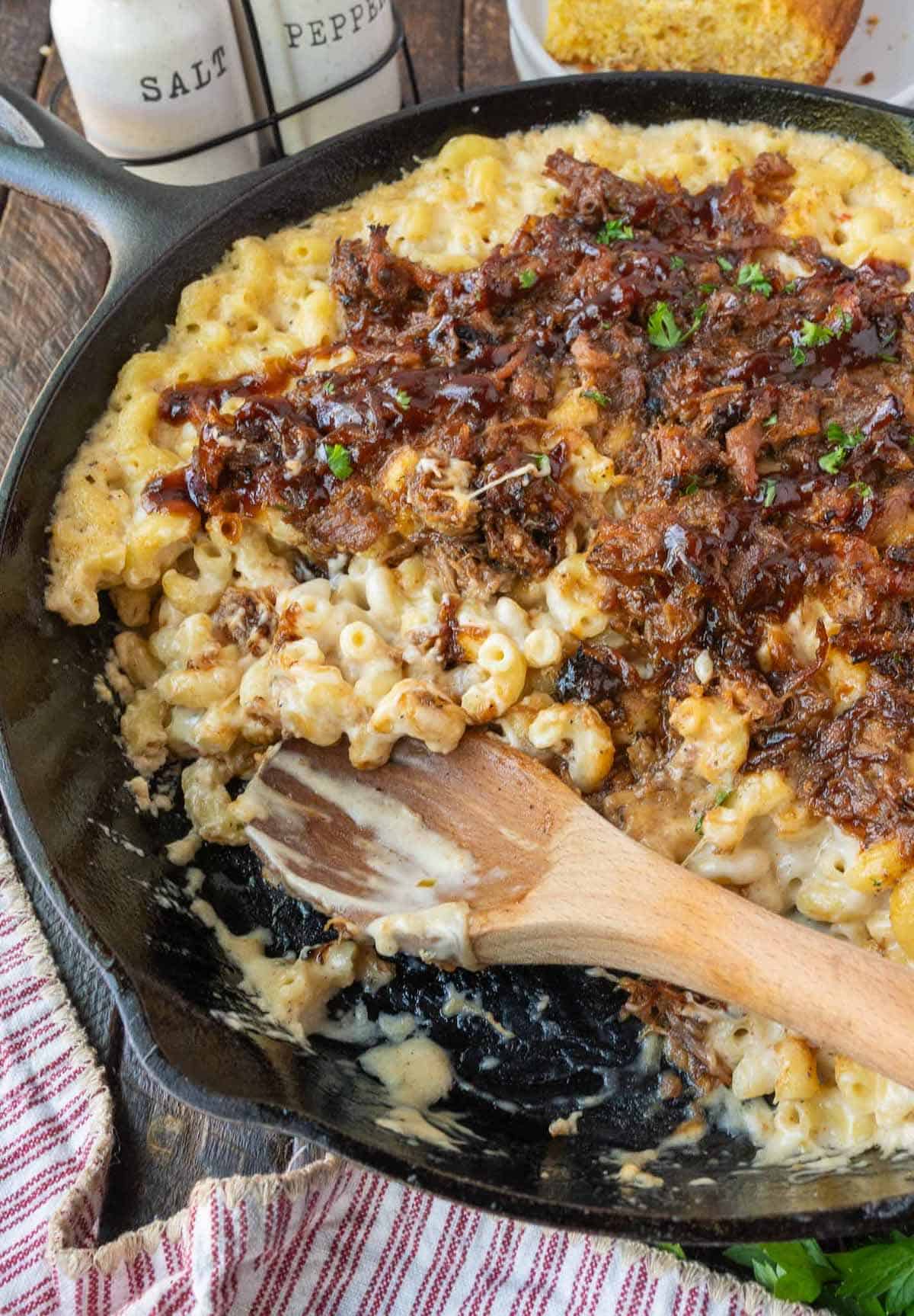 A wooden spoon in a cast iron skillet with pulled pork mac and cheese.