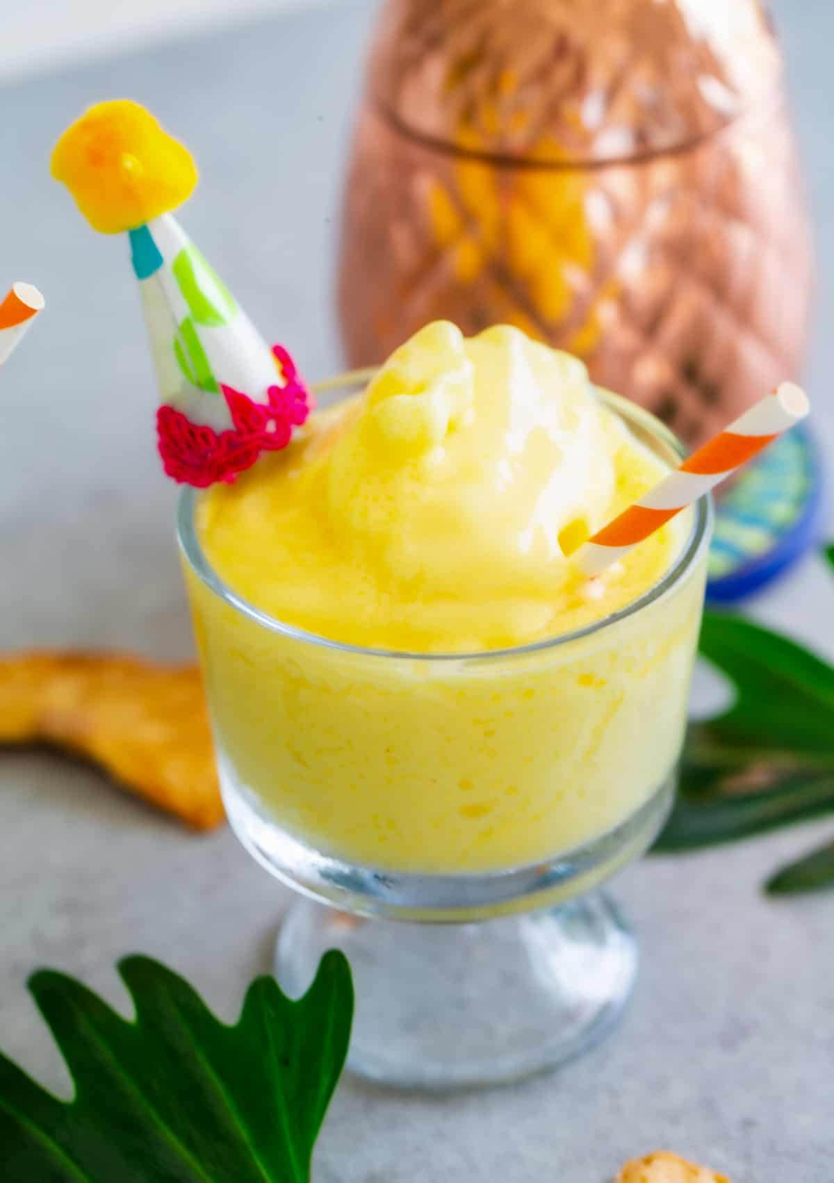 Homemade pineapple dole whip in a glass with a decorative party hat and a red striped straw.