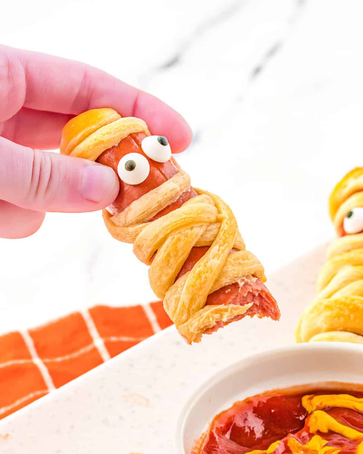 A mummy hot dog with a bite pout of it after being dipped in the sauce.
