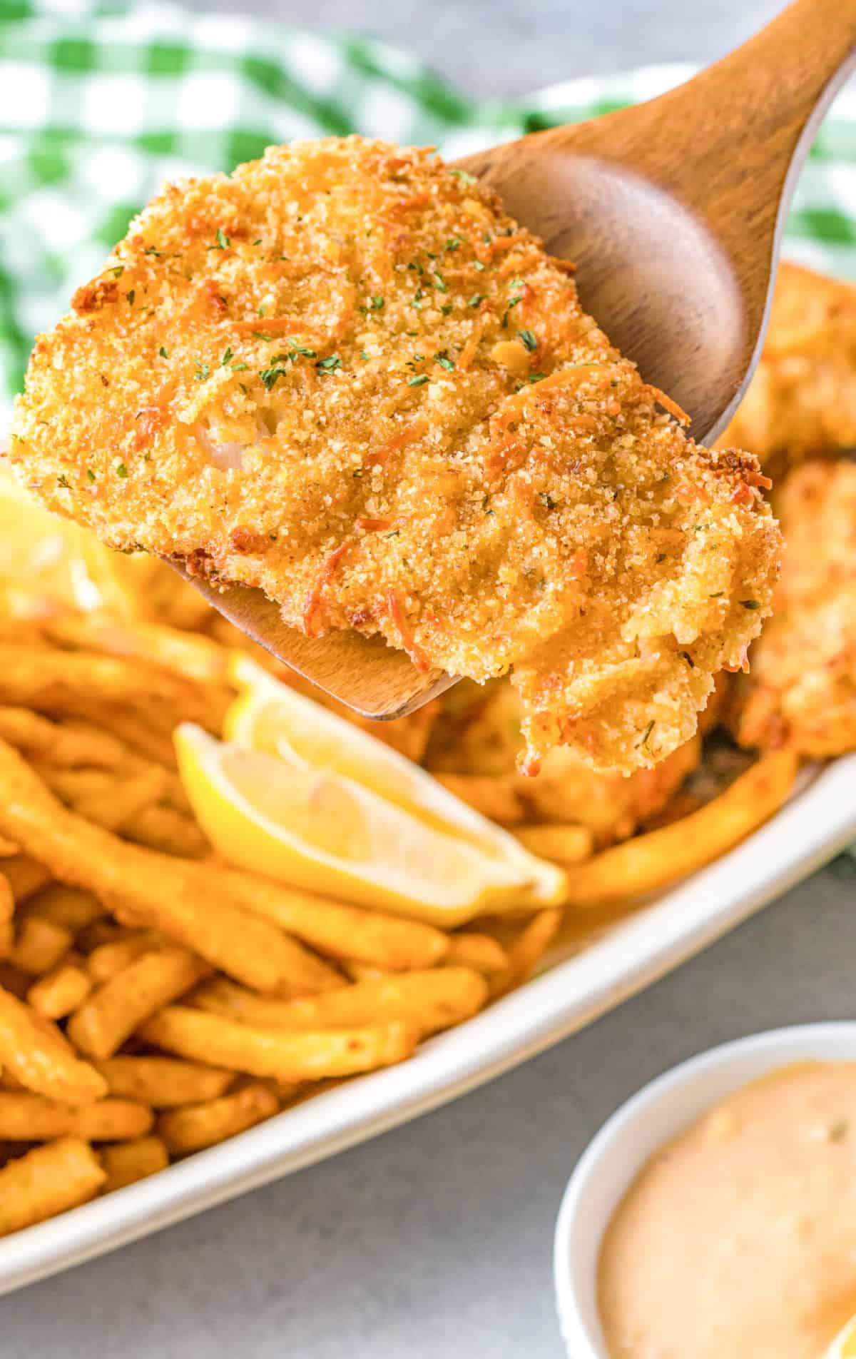 A close up photo of a wooden spatula picking up a breaded fish filet.