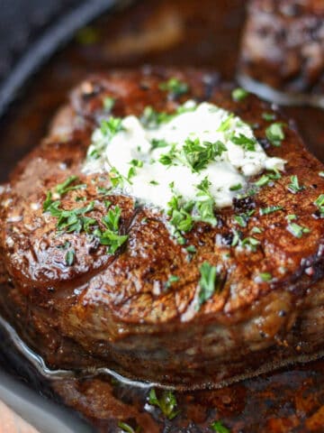 A close up photo of filet mignon steak in a cast iron pan with a dab of garlic herb butter.