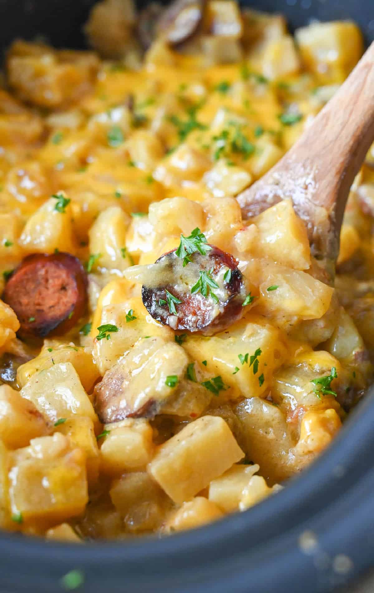 A wooden spoon scooping up some cheesy potatoes with sausage.