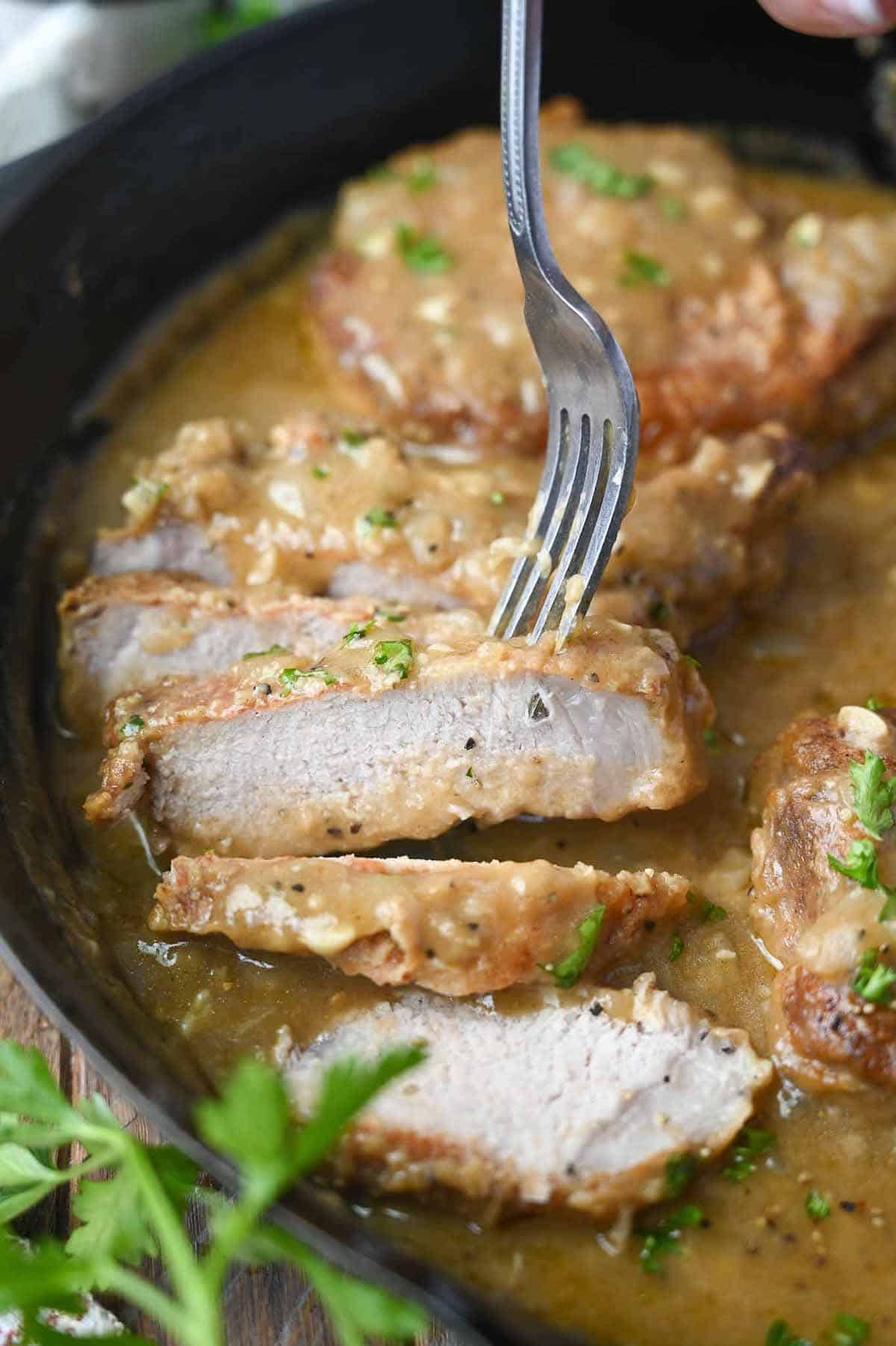 Smothered pork chops in a skillet sliced and a fork pickinh up a bite.