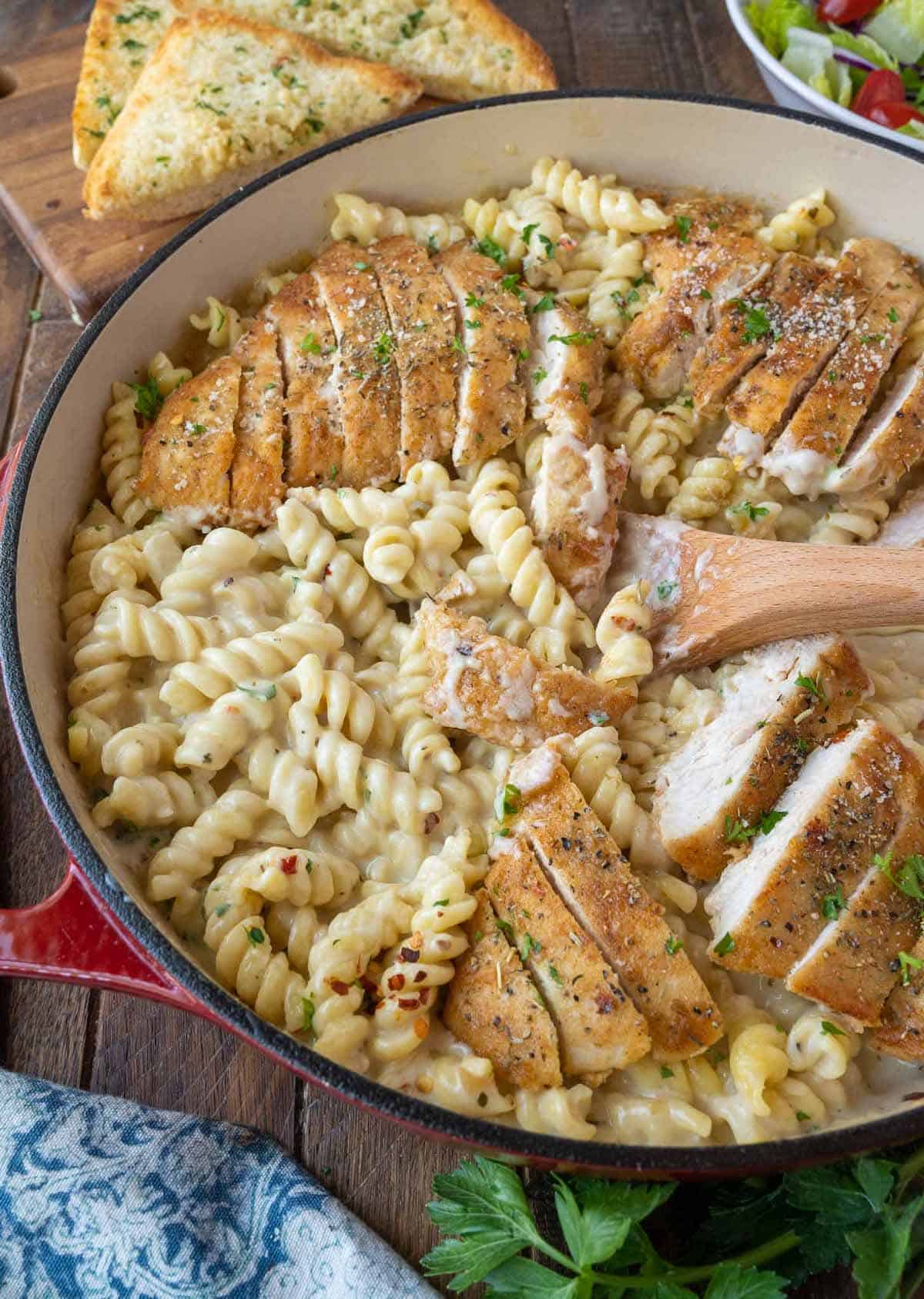 Creamy pasta in a skillet with chicken and a wooden spoon.