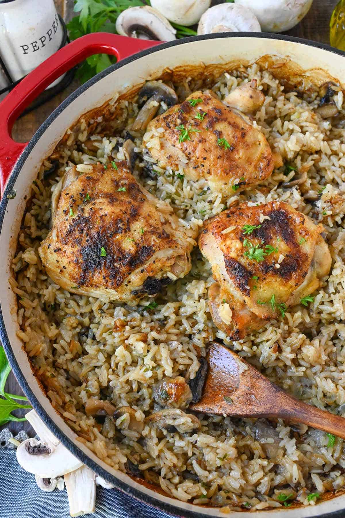 A wooden spoon sitting in a skillet of mushroom rice and chicken thighs.