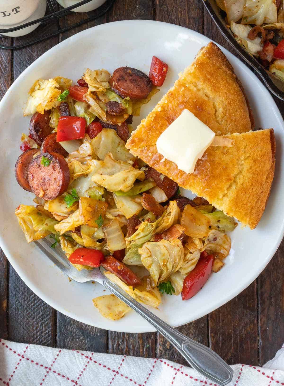 Fried cabbage and sausage on a white plate with a side of cornbread.