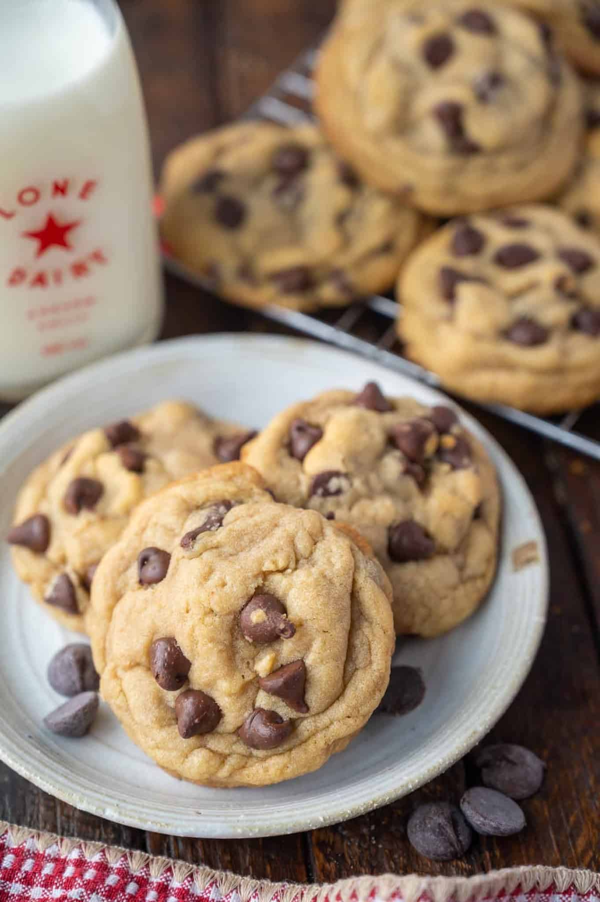 Chocolate chip cookies on a plate with more cookies cooling on a wire rack in the background.