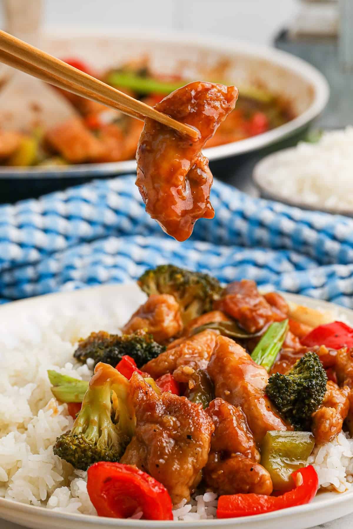 A pair of chopsticks lifting a piece of chicken above a plate of spicy hunan chicken.