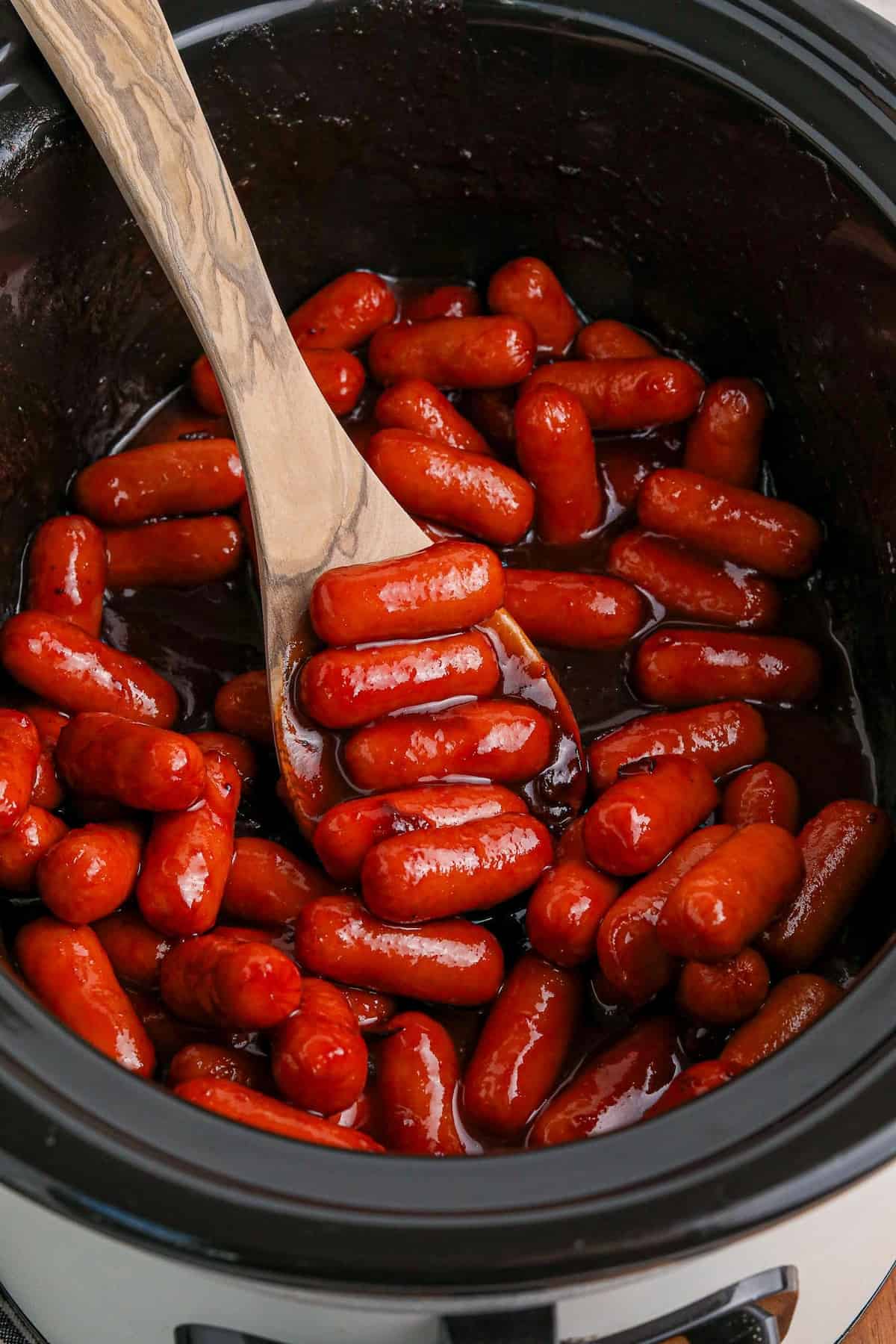 Stirring slow cooker cranberry little smokies with a wooden spoon.