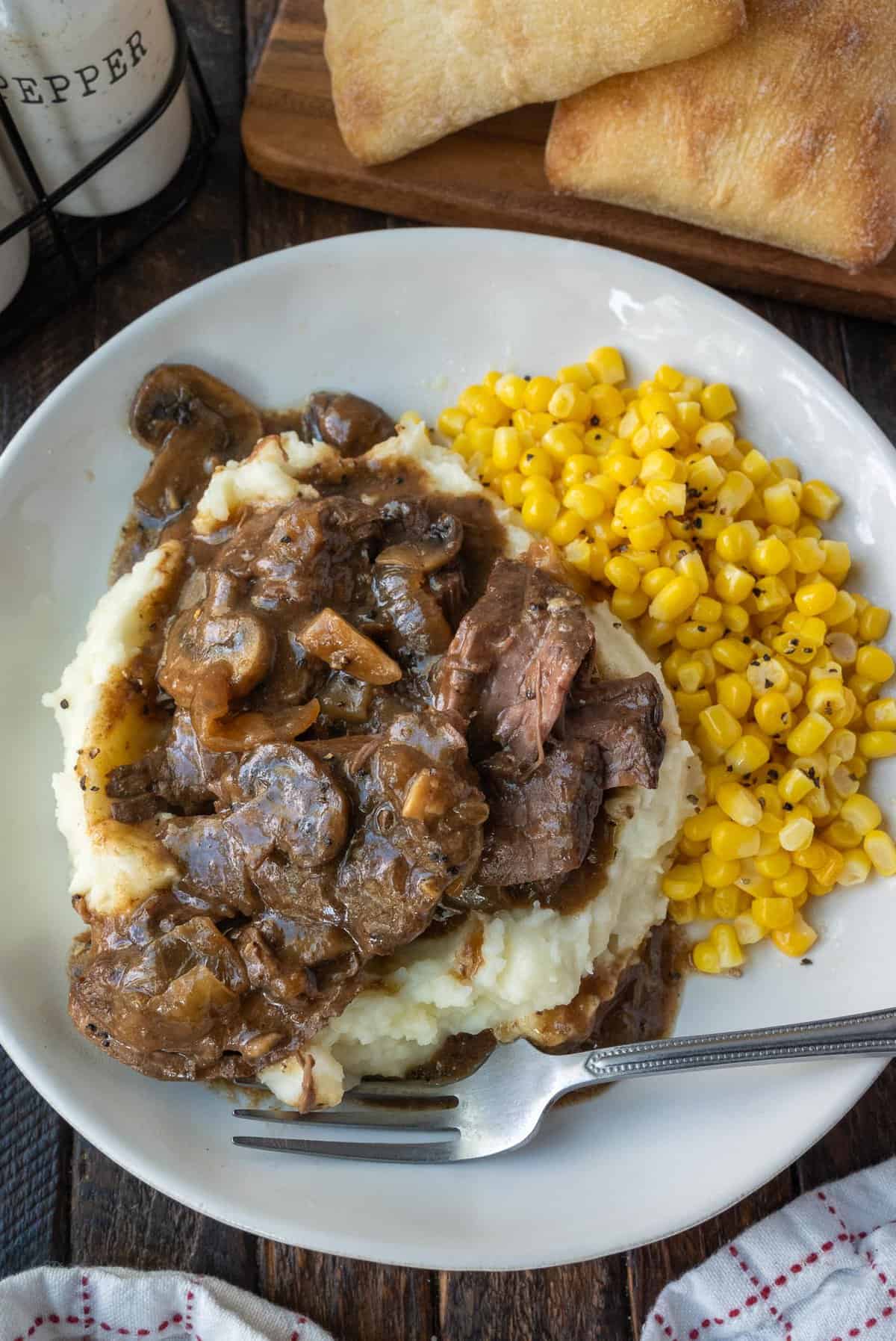 Sliced steak in gravy on top of mashed potatoes with a side of corn.