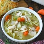 A bowl of turkey noodle soup on a table with spoons and a piece of toasted bread.
