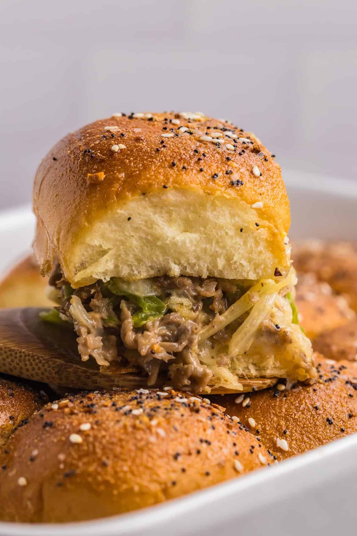 A single cheesesteak slider resting on top of a dish of other baked sliders.