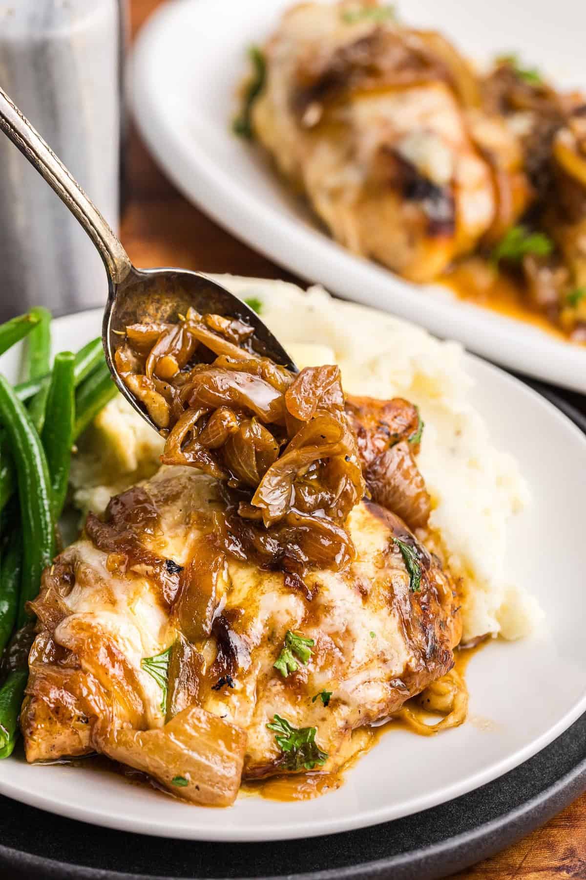 Spooning onions over a plate of french onion chicken bake with mashed potatoes and green beans.