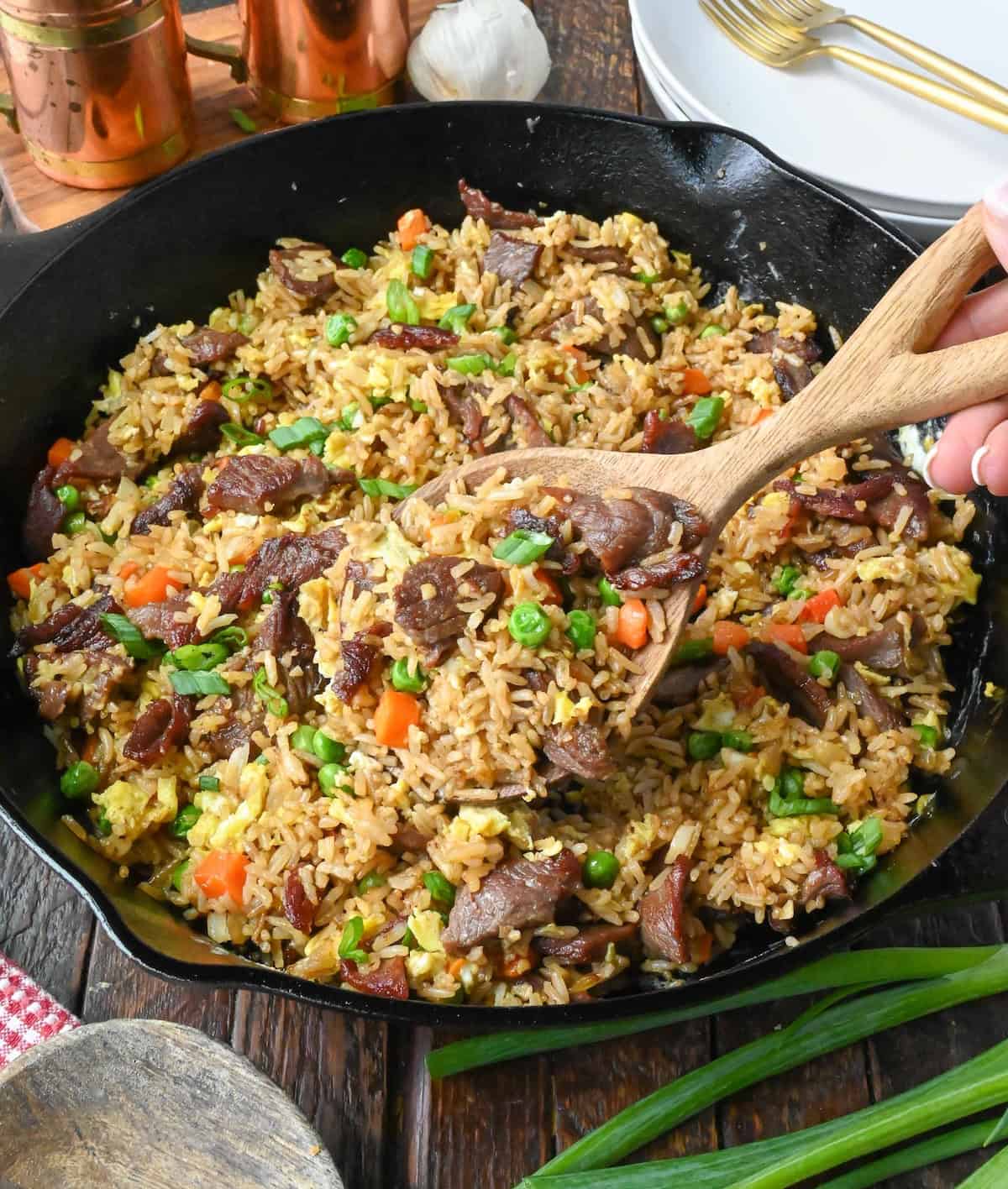 Using a wooden spoon to scoop steak fried rice out of a cast iron skillet.