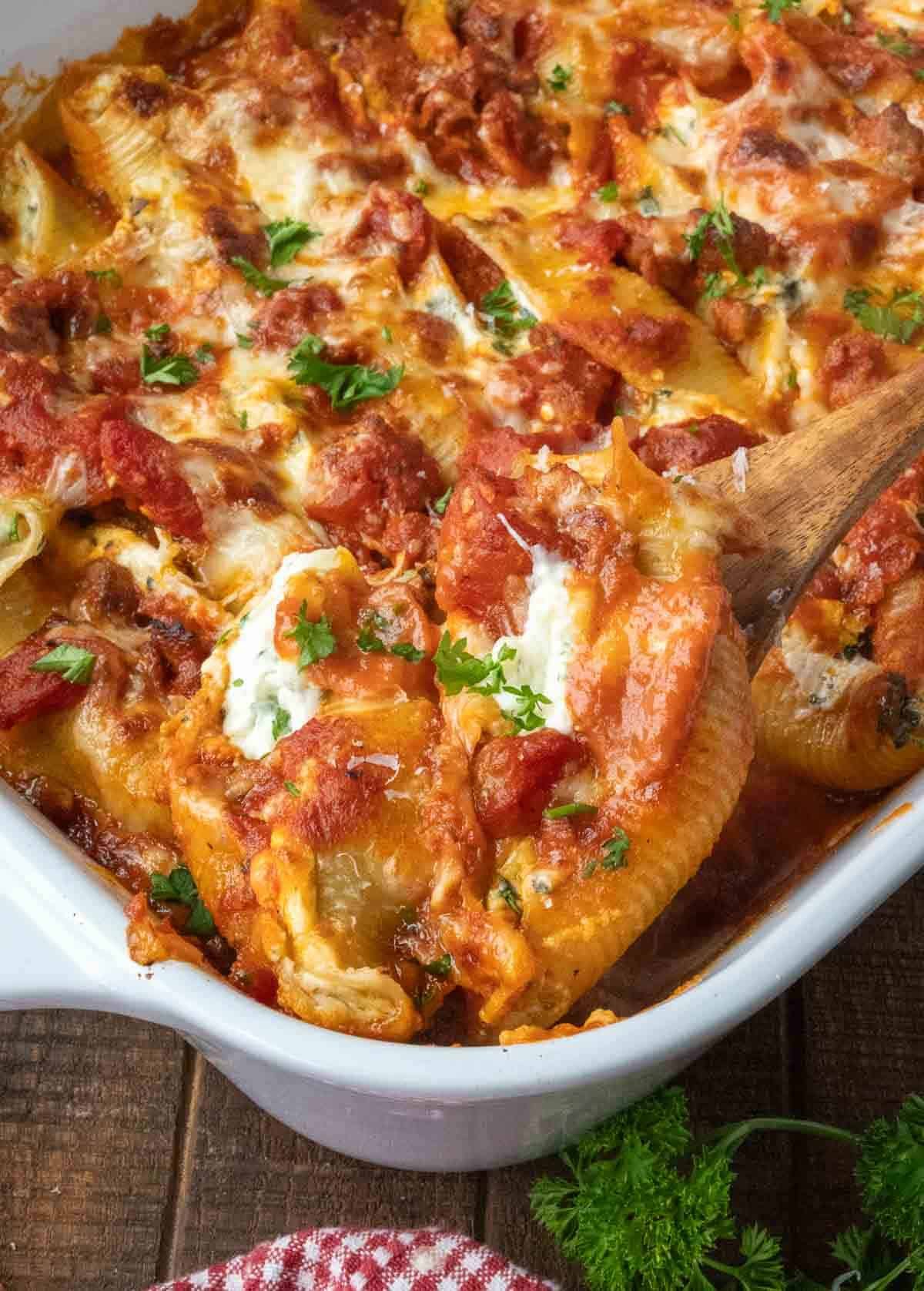 Taking a scoop of cheese stuffed shells from a casserole dish with a wooden spoon.