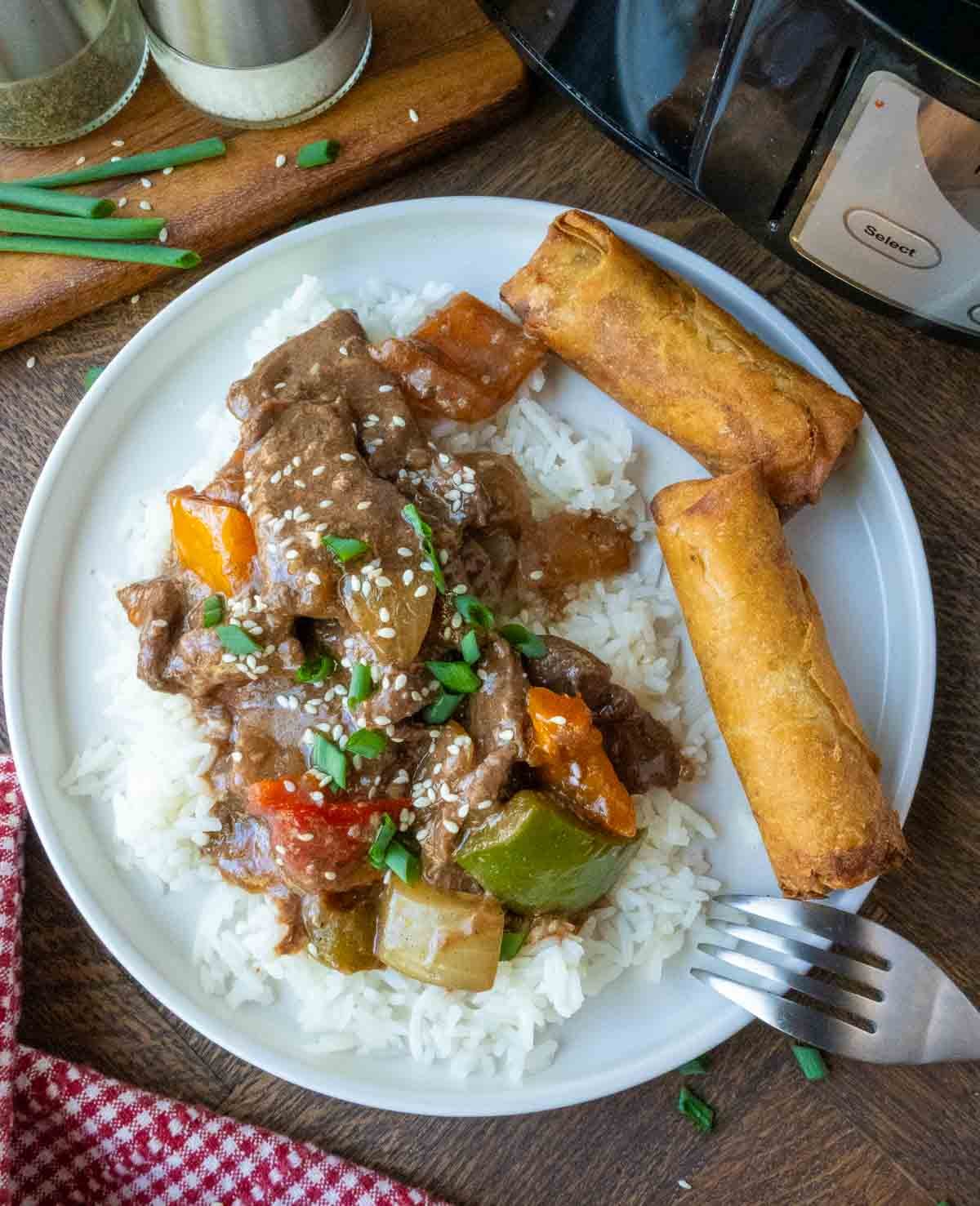 Pepper steak on a plate with white rice and egg rolls.