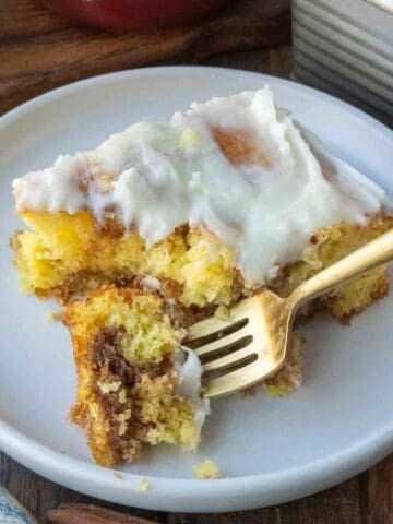 A plate with a golden fork and a slice of cinnamon roll cake with frosting.