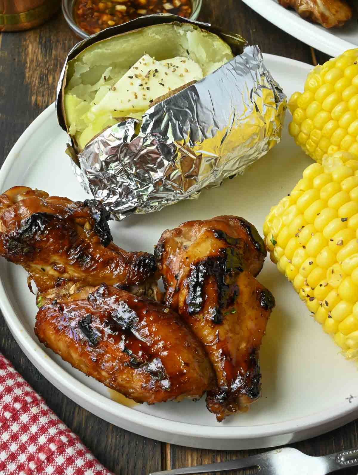 A plate with grilled garlic chicken wings, a baked potato, and corn on the cob.