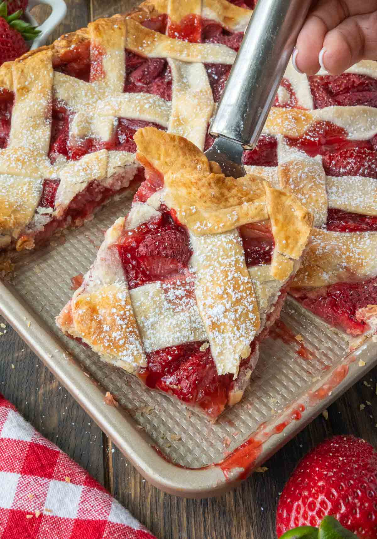 Using a spatula to take a slice of strawberry slab pie from a baking sheet.