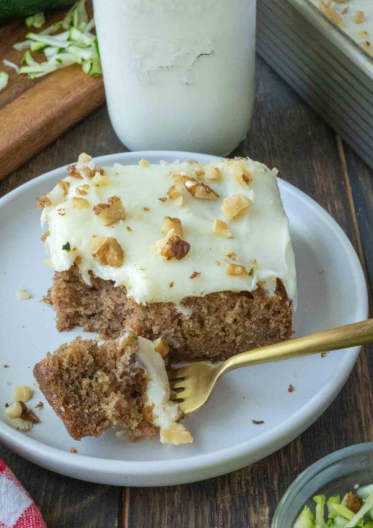 A slice of zucchini cake with frosting on a plate with a gold fork.