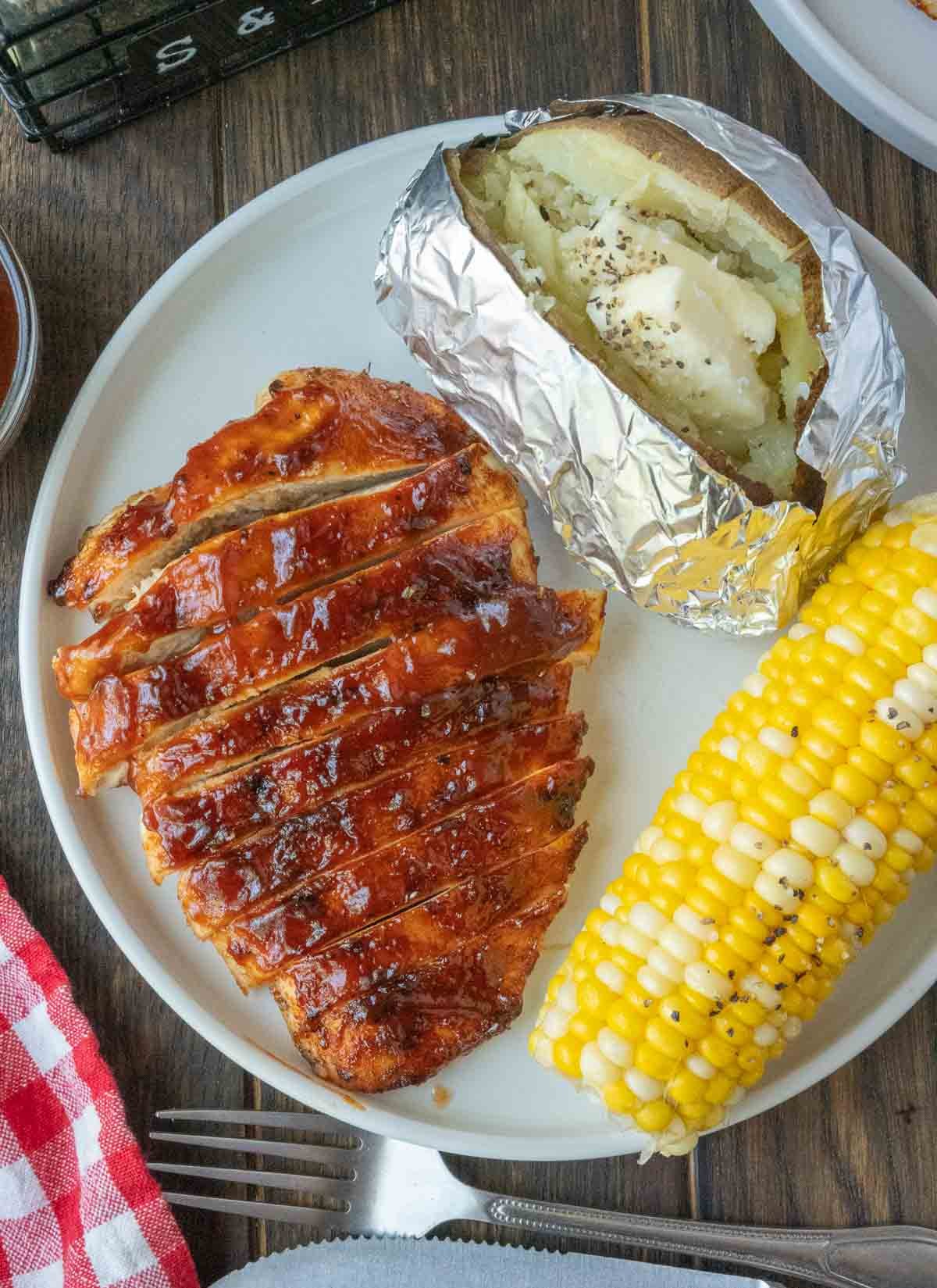 Sliced BBQ chicken on a plate with corn and a potato.