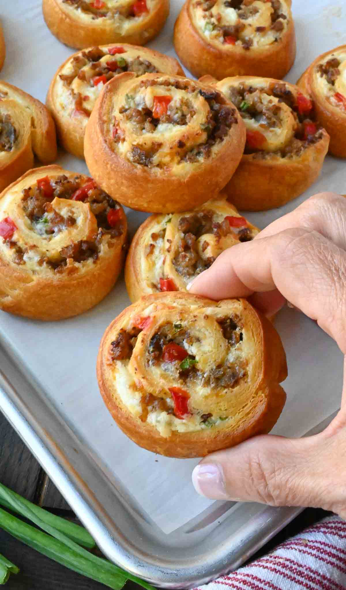Holding up a sausage pinwheel over a tray of more golden brown pinwheels.