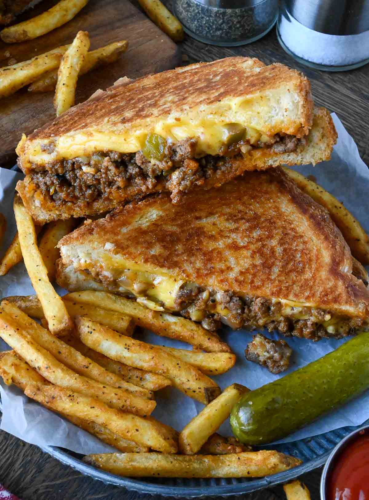 Sloppy joes grilled cheese cut in half on a plate with fries and a pickle.