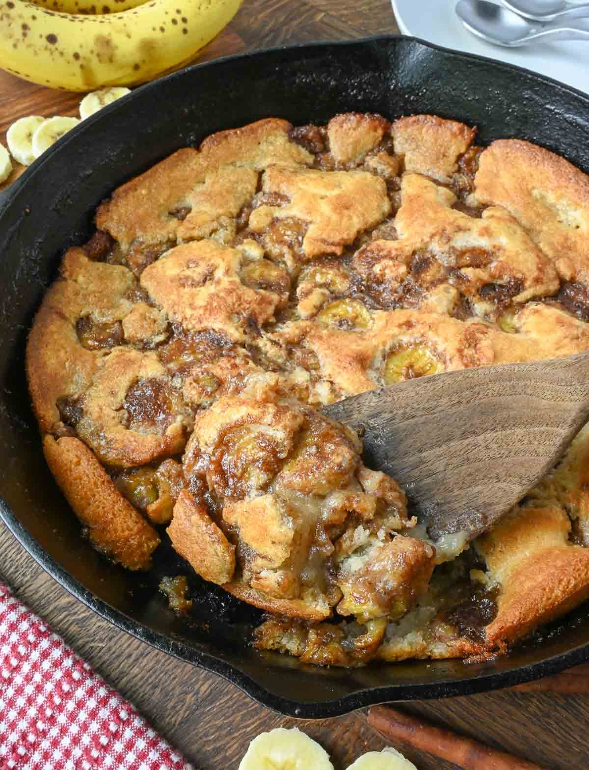Using a wooden spoon to take a scoop of banana cobbler from a cast iron skillet.