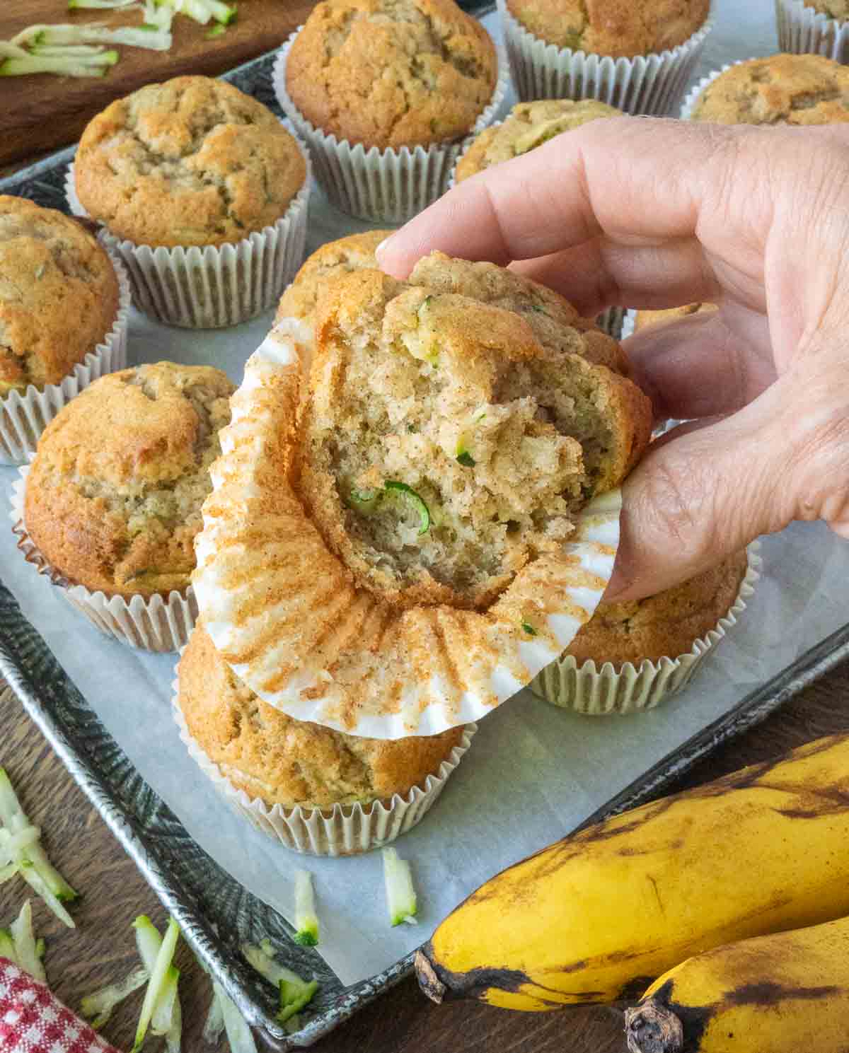 Holding up half of a banana and zucchini muffin over a baking tray full of muffins.