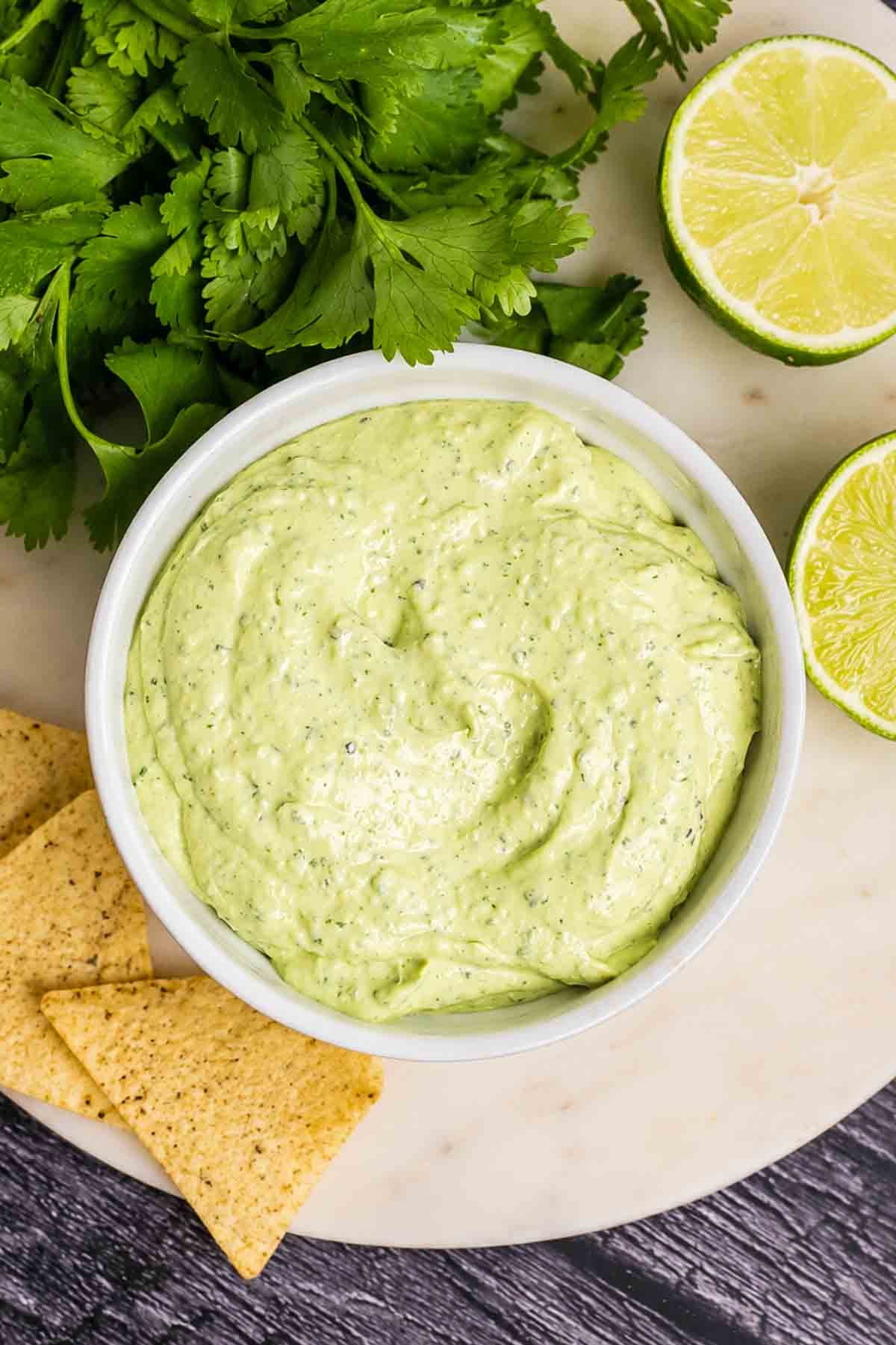 A bowl of creamy avocado sauce with tortilla chips and cilantro on the side.