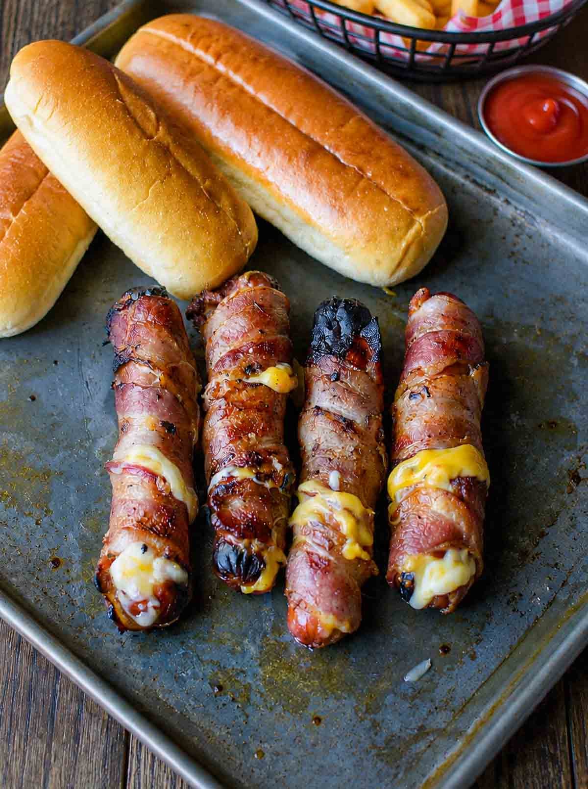 A griddle with 4 bacon wrapped hot dogs and several hot dog buns.