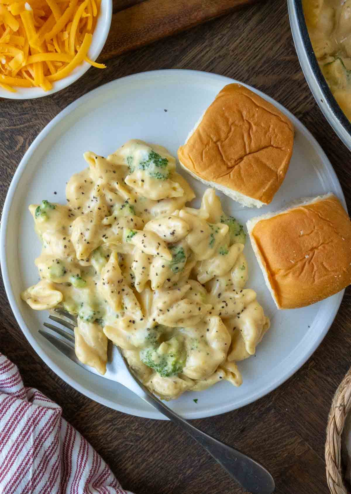 A plate of broccoli mac and cheese served with a side of 2 rolls.