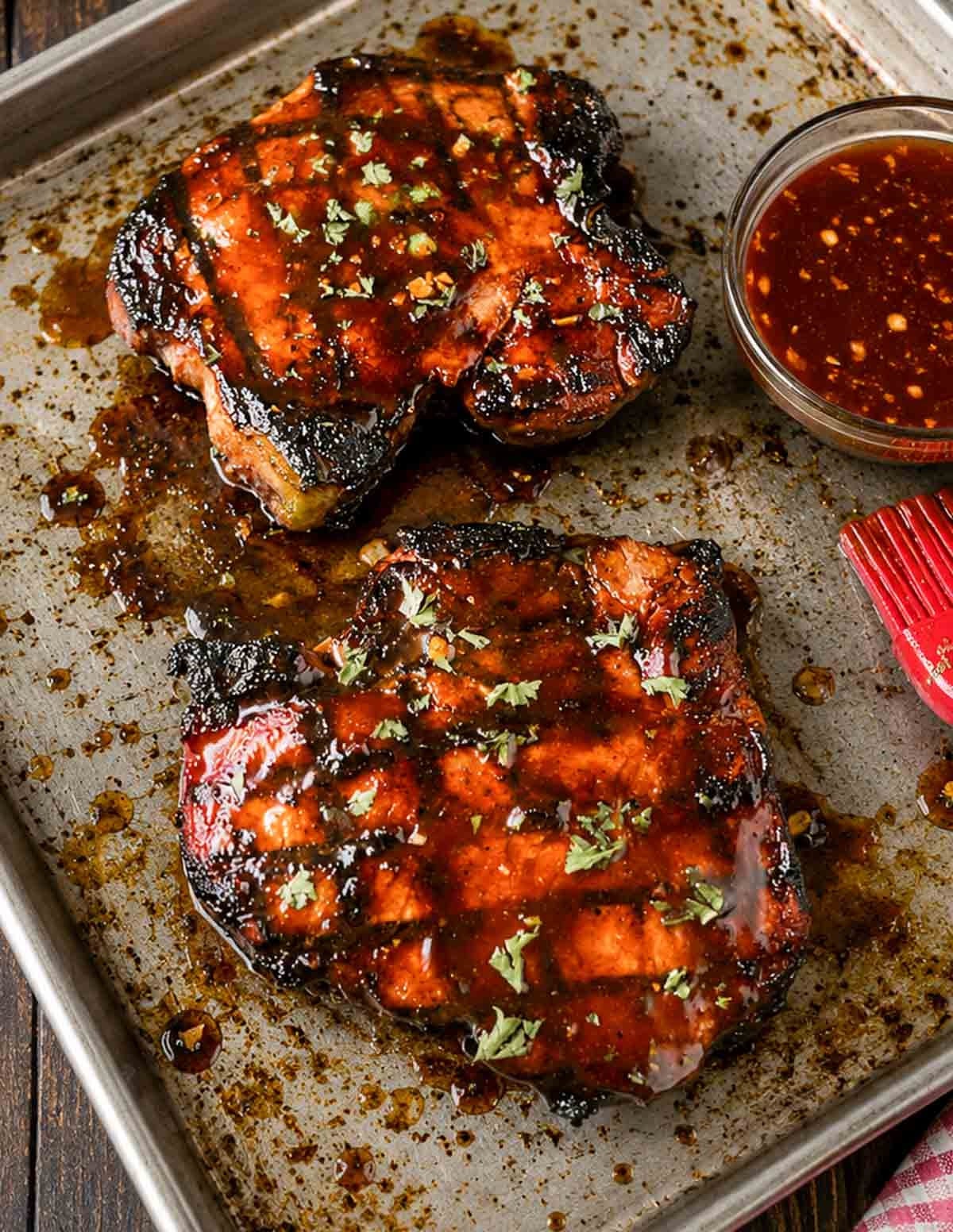 Grilled hoisin pork chops on a sheet pan with extra sauce in a bowl.