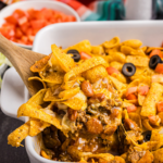A wooden spoon scooping out some frito pie from a dish.